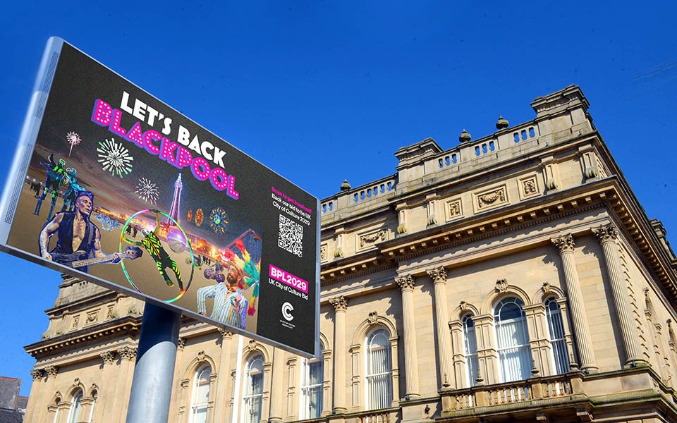 Large digital billboard promoting “LET’S BACK BLACKPOOL” with colourful illustrations of performers, fireworks, and entertainment attractions. The billboard stands in front of a historic stone building with columns and arched windows under a clear blue sky. Visible text includes a QR code and the slogan encouraging support for Blackpool’s bid to become the UK City of Culture 2029.