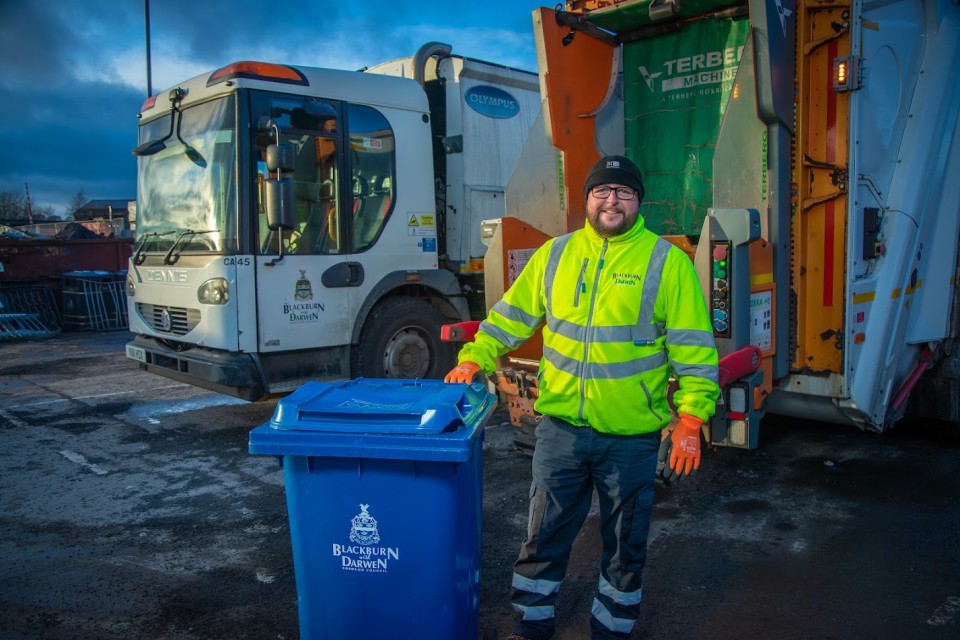 Waste collector in hi vis jacket standing next to blue recycling bin.