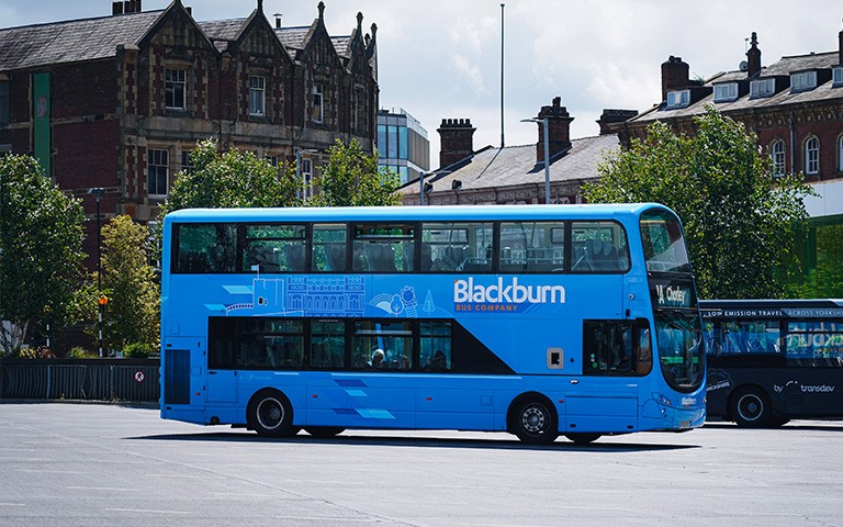 A bright blue double-decker Blackburn Bus Company bus is parked in an open bus station area, with trees and historic brick buildings in the background. The side of the bus displays the Blackburn Bus Company branding and local landmark illustrations, while another bus is partially visible to the right. The scene is captured in daylight under a partly cloudy sky.