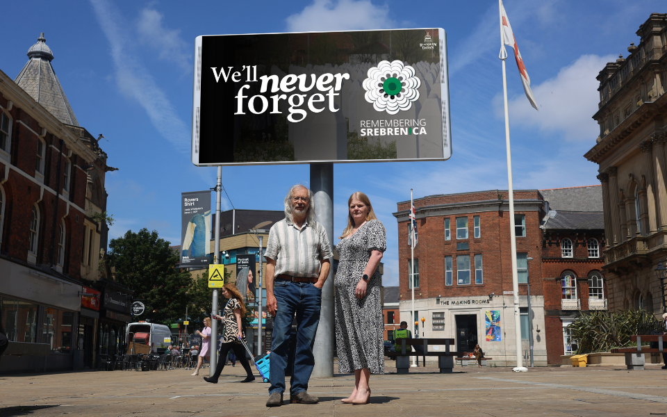 Phil and Denise standing in front of the big screen with the words we will never forget